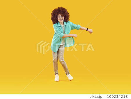 Happy cheerful playful boy in funny red curly wig, dressed in big blue shirt and gray jeans, is fooling around and dancing in good mood. 100234218