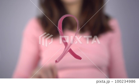 A young woman paints a breast cancer symbol on a glass. The girl draws a pink loop on the glass with a brush. 100234986