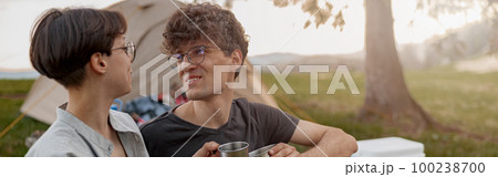 Young male and female sitting at nature near tent, talking and drinking tea or coffee. Young male and female sitting at nature near tent, talking and drinking tea or coffee. 100238700