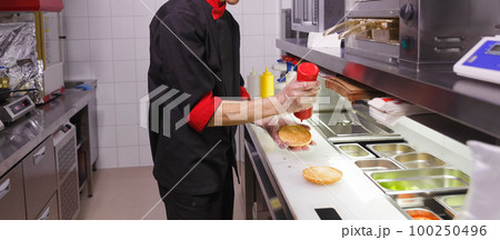 Cook adding a ketchup on burger. 100250496