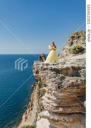 Woman in a yellow dress on the sea. Side view Young beautiful sensual woman in yellow long dress posing on a rock high above the sea at sunset. Girl in nature against the blue sky Woman in a yellow dress on the sea. Side view Young beautiful sensual woman in yellow long dress posing on a rock high above the sea at sunset. Girl in nature against the blue sky 100256885