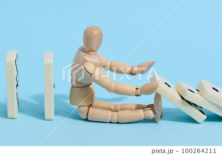 A wooden puppet toy holds back a falling domino on a blue background, representing the concept of a strong personality 100264211