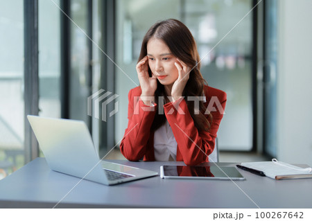 A portrait of a beautiful Asian female employee showing a stressed face while using the phone and financial documents on her desk. 100267642