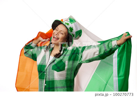 Happy woman dressed in green checkered shirt and carnival hat with clover leaves, smiling and carrying Irish flag, celebrating Saint Patrick's Day on isolated white background. Culture and traditions 100270773