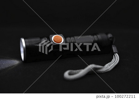 Metal black flashlight on a black background. Burning pocket flashlight close-up. Lighting device. 100272211