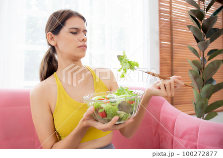 Young caucasian woman sitting on sofa eating vegetable salad while feeling dislike. Young caucasian woman sitting on sofa eating vegetable salad while feeling dislike. 100272877