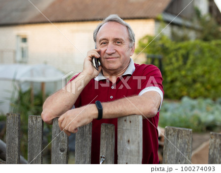 Mature man standing relaxed near fence talking on phone in backyard smiling Mature man standing relaxed near fence talking on phone in backyard smiling 100274093