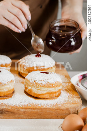 Woman chef prepares fresh donuts with jam at her bakery. Cooking traditional Hanukkah sufganiyot. Small business. 100282564