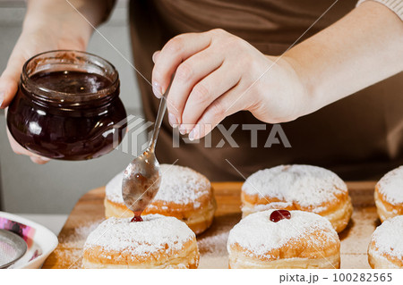Woman chef prepares fresh donuts with jam at her bakery. Cooking traditional Hanukkah sufganiyot. Small business. 100282565