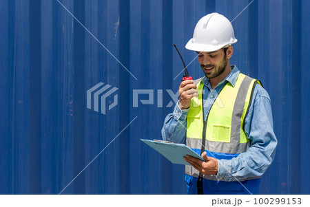 Shipment worker with safety vest and hardhat looking at clipboard  while holding walkie talkie. A large steel cargo container is in the background. 100299153