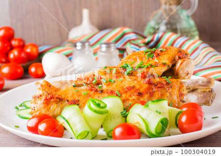 Baked in a spices turkey wing, cucumber slices and cherry tomatoes on a plate on a wooden table 100304019
