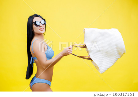 Young sexy woman slim body wearing in blue swimsuit and with sunglasses holding big shopper bag, walking on isolated on yellow color background studio. Summer rest concept 100317351