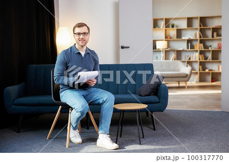 Portrait of happy male psychologist looking at camera and taking notes during therapy session at clinic. Friendly psychotherapist posing and smiling at office 100317770