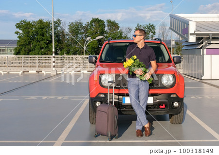 A young man with a bouquet of flowers and a suitcase stands outside his car outdoors A young man with a bouquet of flowers and a suitcase stands outside his car outdoors 100318149