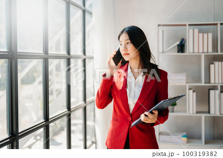 Business asian woman Talking on the phone and using a laptop with a smile while sitting at office Business asian woman Talking on the phone and using a laptop with a smile while sitting at office 100319382