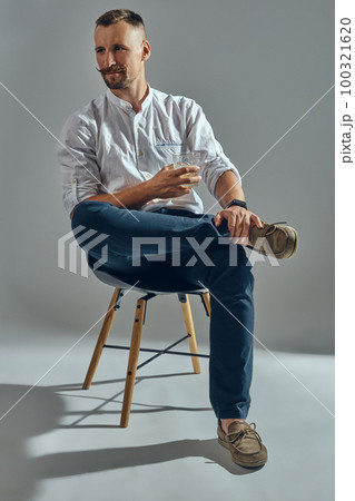 Man with stylish mustache, in classic white shirt and blue trousers is sitting on chair, holding glass of whiskey. Grey background, close-up shot. Man with stylish mustache, in classic white shirt and blue trousers is sitting on chair, holding glass of whiskey. Grey background, close-up shot. 100321620