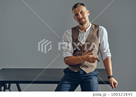 Man with a stylish mustache, dressed in brown vest, white shirt and dark trousers is sitting on table, enjoying cigar. Grey background, close-up. Man with a stylish mustache, dressed in brown vest, white shirt and dark trousers is sitting on table, enjoying cigar. Grey background, close-up. 100321645