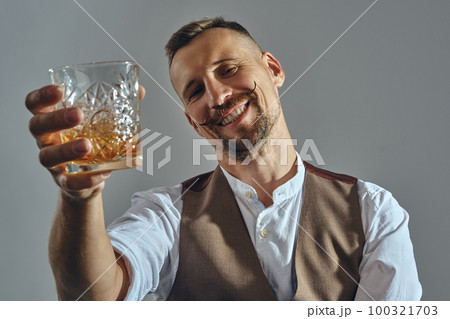 Man with stylish mustache, dressed in classic brown vest, white shirt is sitting at the table, enjoying whiskey. Grey background, close-up shot. Man with stylish mustache, dressed in classic brown vest, white shirt is sitting at the table, enjoying whiskey. Grey background, close-up shot. 100321703