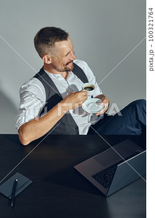 Businessman with stylish mustache, dressed in classic suit is smiling, looking away while sitting at table in office and having coffee. Top view. 100321764