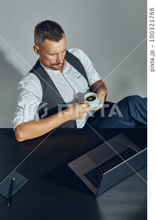 Bearded businessman with stylish mustache, dressed in classic suit is looking at laptop while sitting at table in office and having coffee. Top view. Bearded businessman with stylish mustache, dressed in classic suit is looking at laptop while sitting at table in office and having coffee. Top view. 100321766