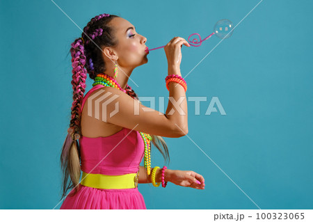 Lovely girl with a multi-colored braids hairstyle and bright make-up, is blowing bubbles using a tubule, posing in studio against a blue background. Lovely girl with a multi-colored braids hairstyle and bright make-up, is blowing bubbles using a tubule, posing in studio against a blue background. 100323065