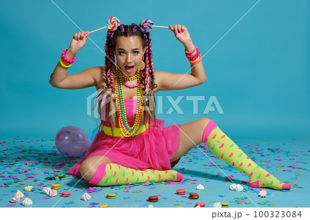 Lovely girl with a multi-colored braids hairstyle and bright make-up, posing in studio with lollipop, air balloons and confetti against a blue background. Lovely girl with a multi-colored braids hairstyle and bright make-up, posing in studio with lollipop, air balloons and confetti against a blue background. 100323084
