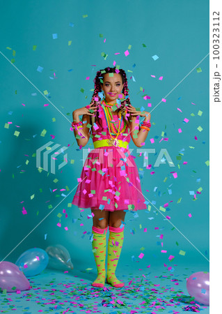 Lovely girl with a multi-colored braids hairstyle and bright make-up, posing in studio with air balloons and confetti against a blue background. 100323112
