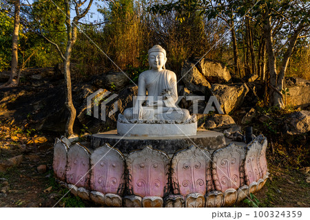 White meditating Buddha statue sits on the rock pile among forest trees in the afternoon time at India. Great religious architecture. 100324359