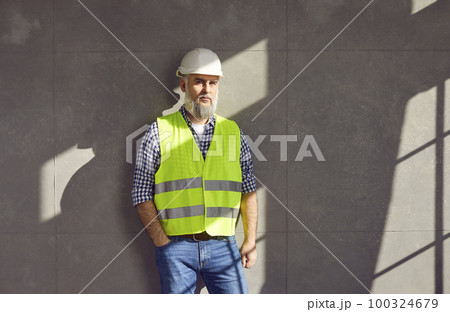 Mature builder or construction engineer in uniform and hard hat standing by grey house wall 100324679