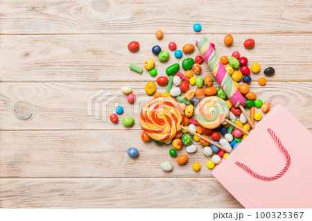 shopping paper gift bag in corner full of assorted traditional candies falling out on colored background with copy space. Happy Holidays sale concept shopping paper gift bag in corner full of assorted traditional candies falling out on colored background with copy space. Happy Holidays sale concept 100325367