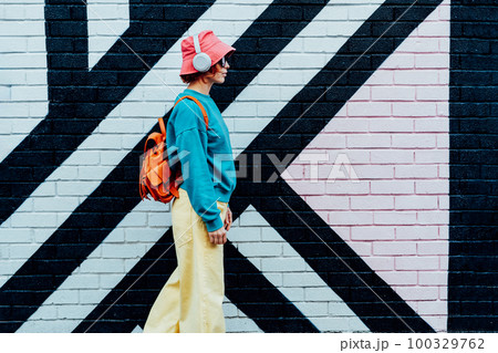 Side view stylish woman in bright clothes and bucket hat wearing wireless headphones and listening to music while walking near city urban painted wall. Fashionable hipster lifestyle. Selective focus. 100329762