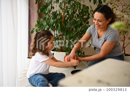 Smiling loving mother and her cute little daughter enjoying floricultural hobby together, watering the soil in pot with planted seeds in the home veranda. Family leisure activity. People and gardening 100329813