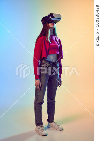 One teen girl wearing VR headset glasses over gradient blue-yellow background. Youth and virtual lifestyle of future. Concept of games, digitalization and technology 100330429