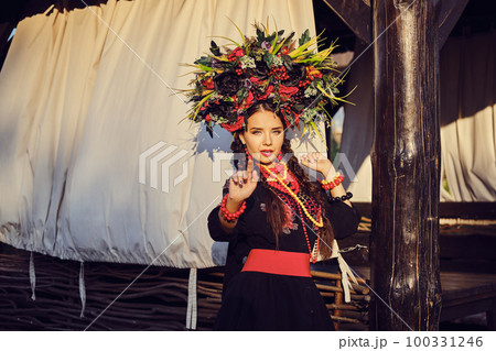 Brunette girl in a white ukrainian authentic national costume and a wreath of flowers is posing against a terrace. 100331246