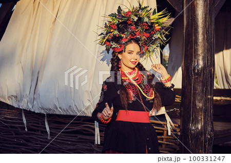 Brunette girl in a white ukrainian authentic national costume and a wreath of flowers is posing against a terrace. 100331247