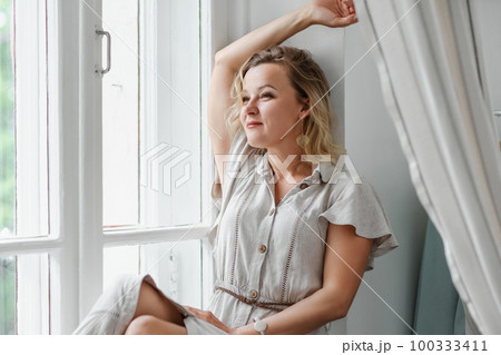 A middle-aged woman in a cream dress sits mysteriously and looks out the window on the windowsill. Green trees outside. 100333411