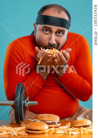 Middle-aged man with beard, dressed in a red turtleneck, headband, posing with burgers and french fries. Blue background. Close-up. Fast food. 100333438