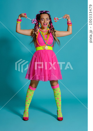 Lovely girl with a multi-colored braids hairstyle and bright make-up, posing in studio against a blue background, holding a lollipop in her hand. Lovely girl with a multi-colored braids hairstyle and bright make-up, posing in studio against a blue background, holding a lollipop in her hand. 100333479