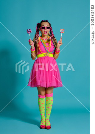 Lovely girl with a multi-colored braids hairstyle and bright make-up, posing in studio against a blue background, holding a lollipop in her hand. Lovely girl with a multi-colored braids hairstyle and bright make-up, posing in studio against a blue background, holding a lollipop in her hand. 100333482