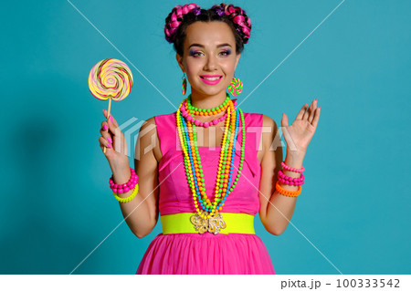 Lovely girl with a multi-colored braids hairstyle and bright make-up, posing in studio against a blue background, holding a lollipop in her hand. 100333542