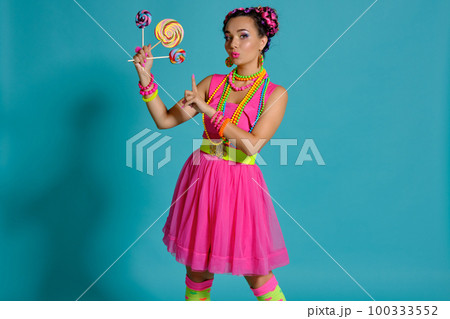 Lovely girl with a multi-colored braids hairstyle and bright make-up, posing in studio against a blue background, holding a lollipop in her hand. Lovely girl with a multi-colored braids hairstyle and bright make-up, posing in studio against a blue background, holding a lollipop in her hand. 100333552