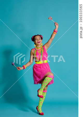 Lovely girl with a multi-colored braids hairstyle and bright make-up, posing in studio against a blue background, holding a lollipop in her hand. Lovely girl with a multi-colored braids hairstyle and bright make-up, posing in studio against a blue background, holding a lollipop in her hand. 100333565