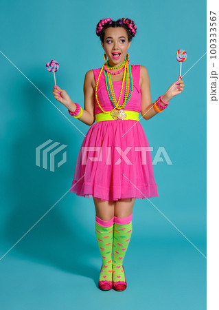 Lovely girl with a multi-colored braids hairstyle and bright make-up, posing in studio against a blue background, holding a lollipop in her hand. Lovely girl with a multi-colored braids hairstyle and bright make-up, posing in studio against a blue background, holding a lollipop in her hand. 100333567