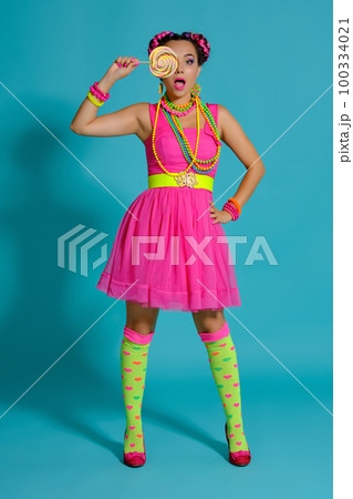 Lovely girl with a multi-colored braids hairstyle and bright make-up, posing in studio against a blue background, holding a lollipop in her hand. Lovely girl with a multi-colored braids hairstyle and bright make-up, posing in studio against a blue background, holding a lollipop in her hand. 100334021