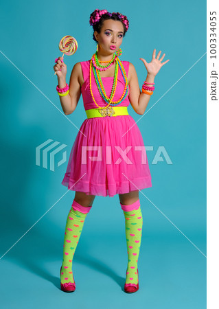 Lovely girl with a multi-colored braids hairstyle and bright make-up, posing in studio against a blue background, holding a lollipop in her hand. 100334055