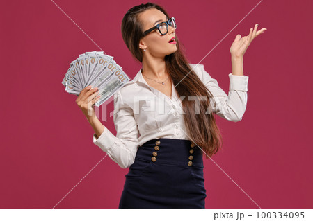 Brunette girl in glasses, wearing in a black short skirt and white blouse is posing holding a fan of hundred dollar bills against a pink background. 100334095
