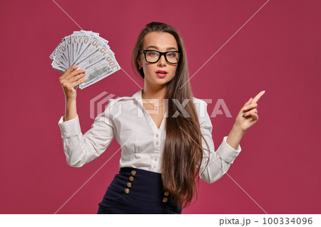 Brunette girl in glasses, wearing in a black short skirt and white blouse is posing holding a fan of hundred dollar bills against a pink background. 100334096