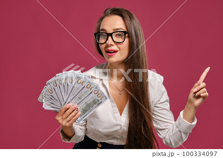 Brunette girl in glasses, wearing in a black short skirt and white blouse is posing holding a fan of hundred dollar bills against a pink background. 100334097