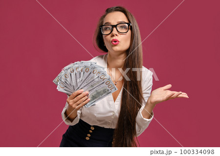 Brunette girl in glasses, wearing in a black short skirt and white blouse is posing holding a fan of hundred dollar bills against a pink background. 100334098