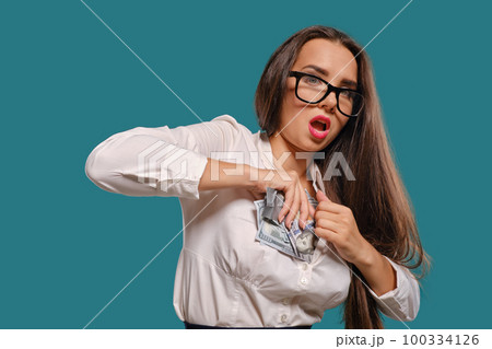 Brunette girl in glasses, wearing in a black short skirt and white blouse is posing holding a fan of hundred dollar bills against a blue background. Brunette girl in glasses, wearing in a black short skirt and white blouse is posing holding a fan of hundred dollar bills against a blue background. 100334126